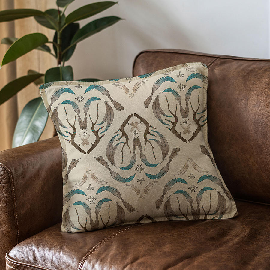 illustration of pattern on cream background of branches crossed in a diamond shape, feathers, bones and runic symbols, tiny skull, gold splatter. Mockup of cushion sitting on the corner of a brown leather sofa.
