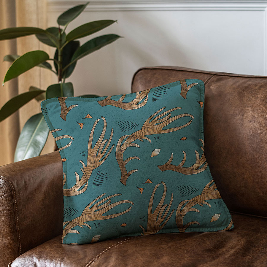 illustration of pattern on teal background of horns, floating teal leaves, citrine crystal, orange crystal. pattern applied on mockup of cushion sitting on corner of leather couch, sofa.